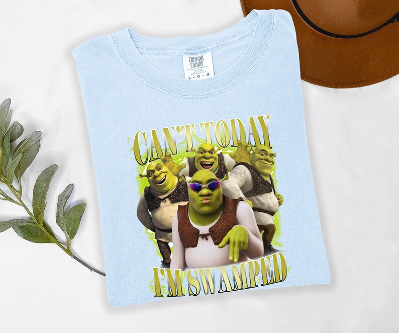May include: Pale blue t-shirt with a graphic of Shrek and the text "CAN'T TODAY I'M SWAMPED" in gold lettering. The image shows Shrek wearing sunglasses with other Shrek characters. The shirt is made of a soft material.