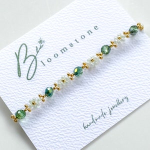 May include: A delicate bracelet with green and white beads and gold accents. The bracelet is on a white card with the text "Bloomstone handmade jewellery".