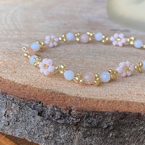Op de afbeelding: Een delicate armband gemaakt met kleine roze en witte kralen, gouden kralen en gouden draad. De armband ligt op een houten oppervlak.