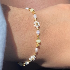 Op de afbeelding: Een witte en gouden kralen armband met kleine witte bloemen bedels. De armband is gemaakt met kleine ronde kralen en gouden spacer kralen.