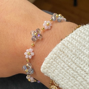 Op de afbeelding: Een delicate armband met een ketting van kleine, witte en paarse bloemvormige kralen afgewisseld met heldere, iriserende kralen. De armband zit om een pols.