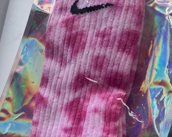 Puede incluir: Calcetines rosas teñidos con un logo Nike negro, empaquetados en una bolsa de plástico transparente e iridiscente. Los calcetines tienen una textura acanalada y un patrón colorido.