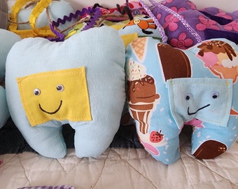 Op de afbeelding: Lichtblauwe, tandvormige pluche speelgoed met gele en bedrukte zakken. Eén heeft een lachend gezicht, de andere heeft een lachend gezicht en een ijsjespatroon. Deze handgemaakte knuffels zijn ontworpen voor kinderen.