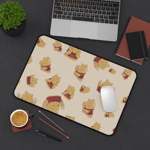 May include: A rectangular mouse pad featuring a repeating pattern of Winnie the Pooh characters in various poses against a light beige background. The mouse pad has a black border and is positioned on a dark surface with a laptop, notebook, and coffee cup.