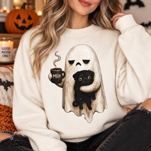 May include: A cream-colored crewneck sweatshirt featuring a graphic illustration of a ghost with a sad expression holding a steaming mug labeled "MEH" and embracing a small black pug dog. The design is centered on the front of the garment.