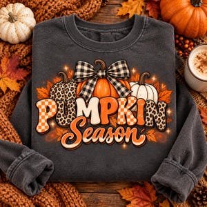 Puede incluir: Una sudadera gris carbón de cuello redondo con un gráfico de tres calabazas y un lazo de cuadros blanco y negro. El texto "PUMPKIN Season" está impreso en naranja y blanco con patrones de cuadros y estampado de leopardo en las letras.