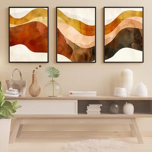 May include: Three framed abstract art prints. Each print features layered watercolour-style designs in shades of brown, orange, and cream, resembling rolling hills. The prints are displayed above a wooden console table with decorative items.