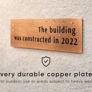 Op de afbeelding: Een rechthoekige koperen plaat met de tekst "The building was constructed in 2022". De plaat is gemonteerd op een getextureerd grijs oppervlak. De tekst "very durable copper plate for outdoor use or areas subject to heavy wear" staat eronder.