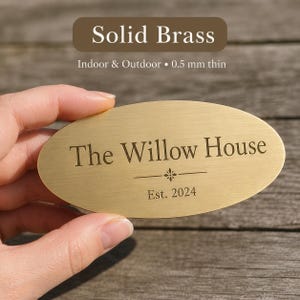 Pode incluir: Placa oval de latão maciço com o texto "The Willow House" e "Est. 2024" em fonte serifada. A placa tem 0,5 mm de espessura e é adequada para uso interno e externo. A placa é segurada por uma mão.