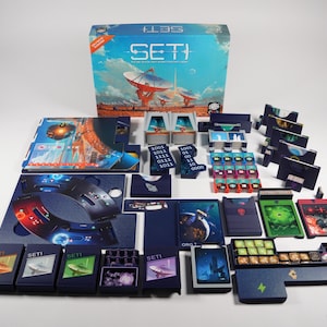 May include: A SETI board game set featuring a large blue box with satellite dish illustrations, multiple dark blue card holders, various game boards, small square tokens with binary code, colorful resource tiles, and miniature game pieces.