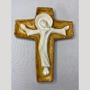 May include: A religious cross with a figure in white against a gold-colored background. The cross has a rectangular shape with rounded edges. The figure is depicted with outstretched arms.