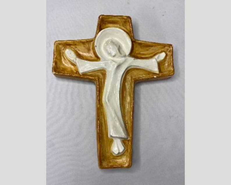 May include: A religious cross with a figure in white against a gold-colored background. The cross has a rectangular shape with rounded edges. The figure is depicted with outstretched arms.