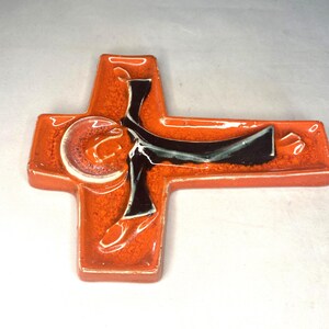 May include: Orange ceramic cross with a black and white figure of a person in a robe. The figure is in a stylized pose with arms outstretched.