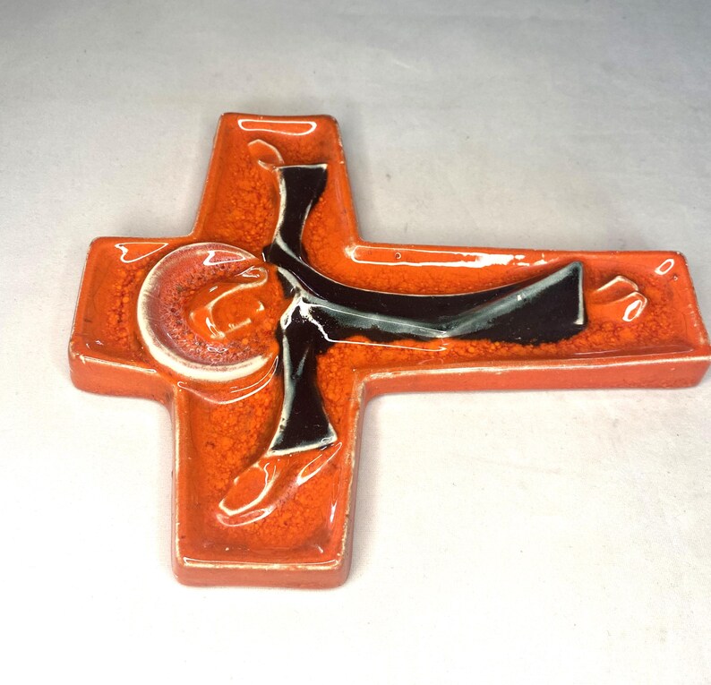 May include: Orange ceramic cross with a black and white figure of a person in a robe. The figure is in a stylized pose with arms outstretched.
