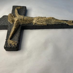 May include: A black ceramic cross with a beige sculpture of a person on it. The sculpture is of a person lying on their back with their arms outstretched.