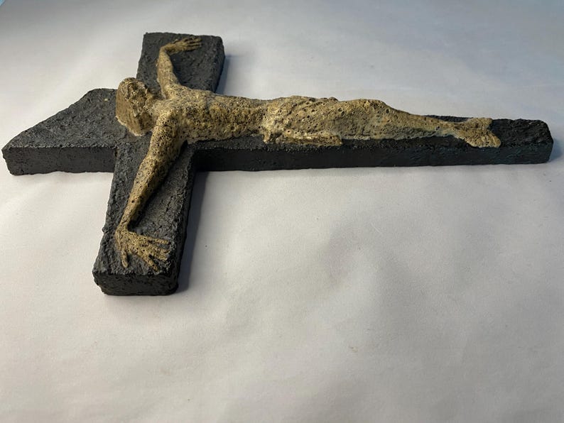 May include: A black ceramic cross with a beige sculpture of a person on it. The sculpture is of a person lying on their back with their arms outstretched.