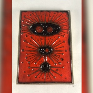 May include: A red ceramic tile with a black and brown abstract design. The design features three stylized flowers with black centers and radiating lines.