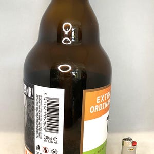 May include: A brown glass beer bottle with a white and black label, and the words "EXTRA ORDINARY" in orange. The bottle contains 330ml of beer and is next to a silver BIC lighter.