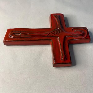May include: A glossy, red ceramic cross with a detailed design. The cross features a textured surface and dark brown accents, creating a striking contrast. The design includes linear and curved elements, adding visual interest to the religious symbol.