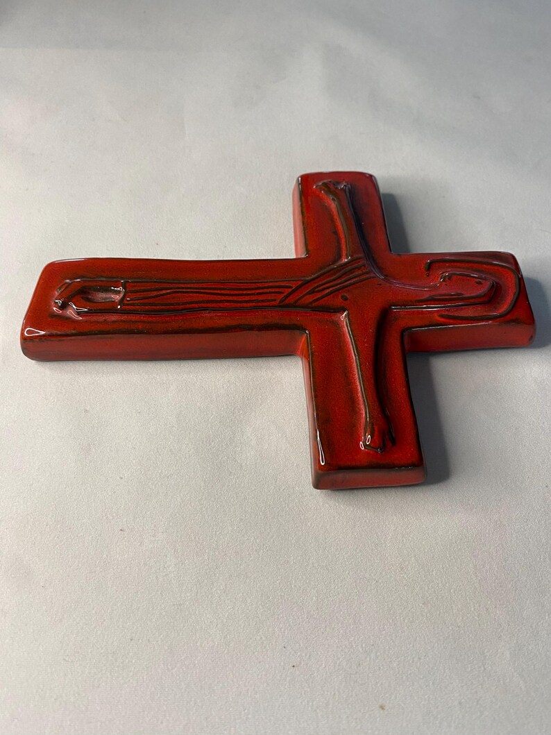May include: A glossy, red ceramic cross with a detailed design. The cross features a textured surface and dark brown accents, creating a striking contrast. The design includes linear and curved elements, adding visual interest to the religious symbol.