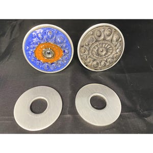 Könnte beinhalten: Vier runde Dekorationsstücke aus Keramik und Metall. Zwei sind silberfarben mit einem erhabenen Augenmotiv. Zwei sind blau mit einem erhabenen orangefarbenen Augenmotiv.