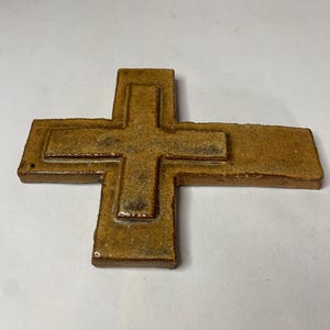 May include: A brown ceramic cross with a raised design. The cross is made of a textured clay and has a rustic look.
