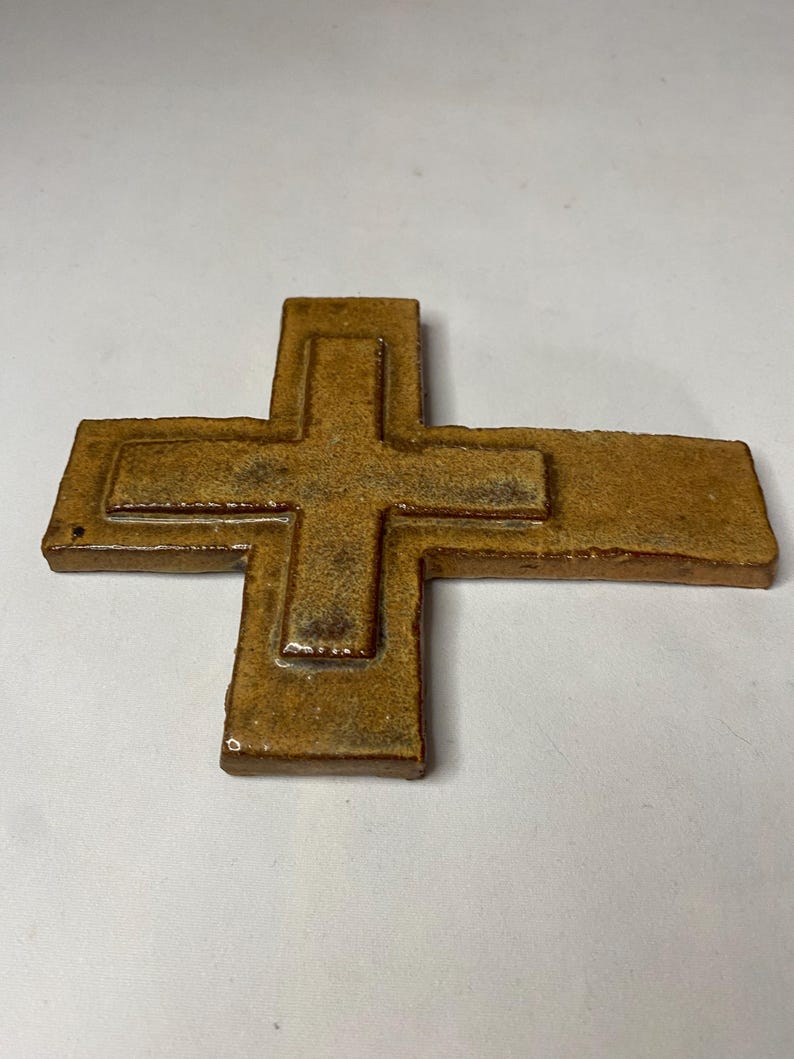 May include: A brown ceramic cross with a raised design. The cross is made of a textured clay and has a rustic look.