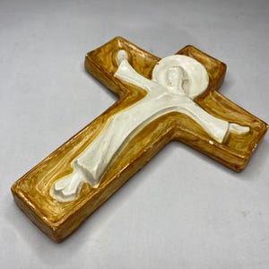 May include: A religious wall cross with a figure in white robes and a halo. The cross is a warm brown color with a raised, textured design. The figure has outstretched arms, and the overall style is simple and reverent.
