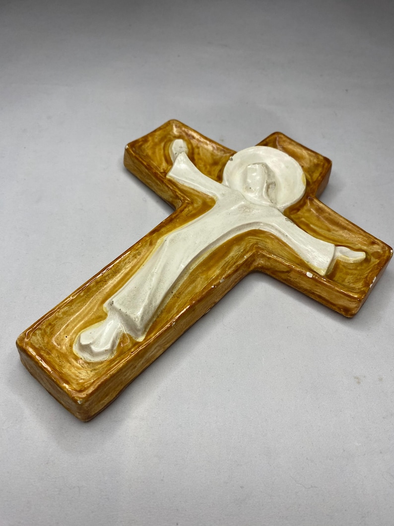 May include: A religious wall cross with a figure in white robes and a halo. The cross is a warm brown color with a raised, textured design. The figure has outstretched arms, and the overall style is simple and reverent.
