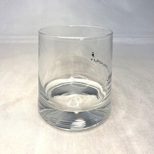 May include: A clear, cylindrical glass with a thick base. The glass features a small black graphic of a person walking and the text "JOHN WALKER" in a vertical, mirrored font. Ideal for serving beverages.