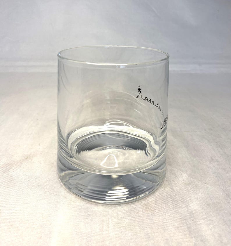 May include: A clear, cylindrical glass with a thick base. The glass features a small black graphic of a person walking and the text "JOHN WALKER" in a vertical, mirrored font. Ideal for serving beverages.