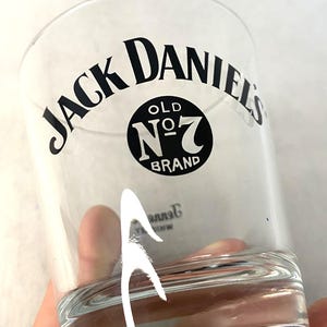 May include: A clear glass tumbler featuring the text "JACK DANIEL'S" in a curved black font. Below the text is a black circle with "OLD NO. 7 BRAND" in white. The glass is held by a hand.