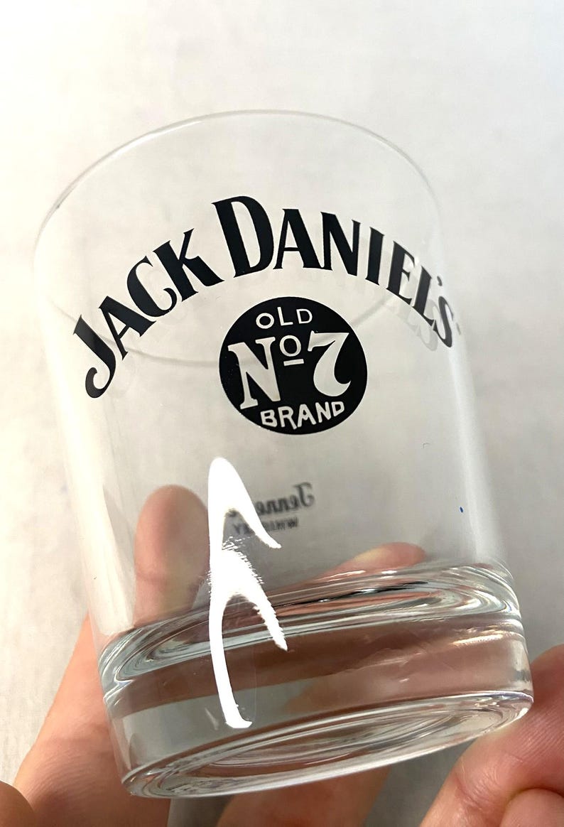 May include: A clear glass tumbler featuring the text "JACK DANIEL'S" in a curved black font. Below the text is a black circle with "OLD NO. 7 BRAND" in white. The glass is held by a hand.