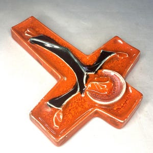 May include: Orange ceramic cross with a black and white figure of a person on it.