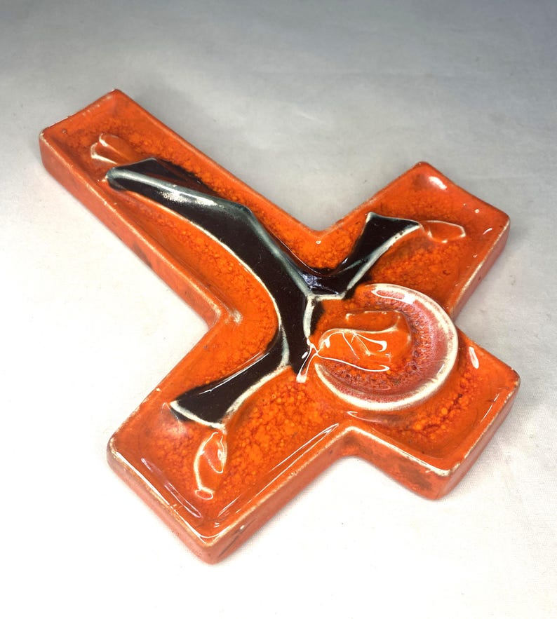 May include: Orange ceramic cross with a black and white figure of a person on it.