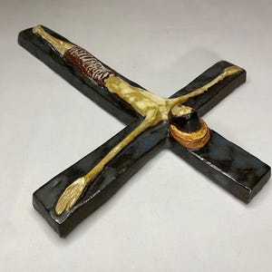 Może przedstawiać: Ceramiczna rzeźba ścienna przedstawiająca krucyfiks z ciemnoniebieską glazurą. Postać Jezusa jest przedstawiona z białym i ż&oacute;łtym ciałem oraz brązowym prześcieradłem. Rzeźba jest zamontowana na czarnym krzyżu.