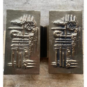 Puede incluir: Dos paneles decorativos rectangulares de color bronce con un diseño abstracto en relieve. Los paneles tienen una superficie texturizada y un acabado metálico oscuro. El diseño parece representar un paisaje urbano o una escena industrial estilizada.
