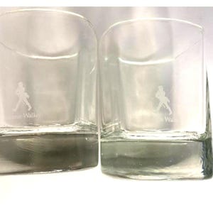 May include: Two clear glass tumblers with a square base and rounded sides. The glasses have a white silhouette of a walking man and the text "Johnnie Walker" etched on the bottom.