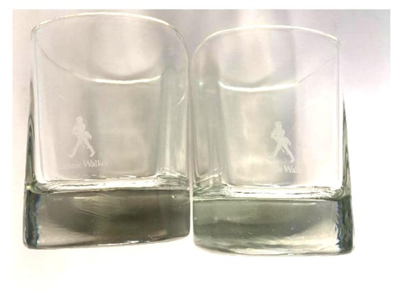 Puede incluir: Dos vasos transparentes de vidrio con base cuadrada y lados redondeados. Los vasos tienen una silueta blanca de un hombre caminando y el texto "Johnnie Walker" grabado en la parte inferior.