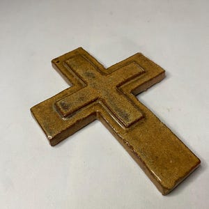May include: A brown ceramic cross with a raised design. The cross has a rustic, aged look.