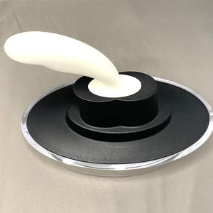 May include: A bottle stopper featuring a white, curved handle and a black base. The base has a circular design with a clear, transparent edge. The stopper is designed to seal bottles.