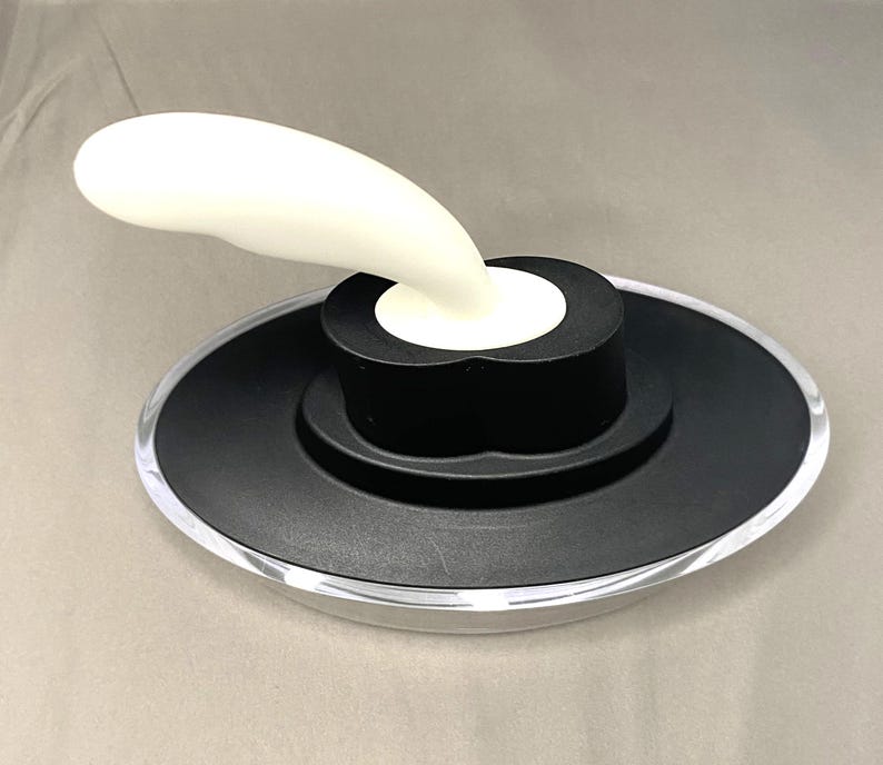 May include: A bottle stopper featuring a white, curved handle and a black base. The base has a circular design with a clear, transparent edge. The stopper is designed to seal bottles.