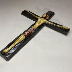Może przedstawiać: Ceramiczny krzyż ścienny z ciemnoniebieską glazurą i beżową figurą Jezusa Chrystusa. Figura jest przedstawiona w brązowym przepasaniu i z rozłożonymi ramionami.