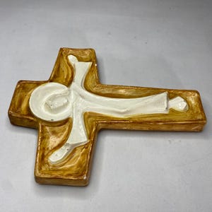 May include: A ceramic cross with a figure in white, set against a brown and cream-colored background. The cross has a rectangular shape with rounded edges. The figure is depicted in a stylized, flowing form.