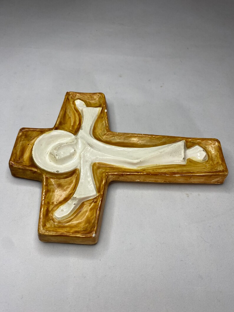 May include: A ceramic cross with a figure in white, set against a brown and cream-colored background. The cross has a rectangular shape with rounded edges. The figure is depicted in a stylized, flowing form.