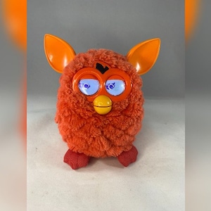 May include: Orange and white Furby toy with blue eyes and a yellow beak. The toy has orange ears and a fluffy orange body.