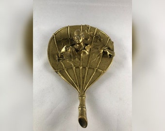 May include: A gold-colored metal fan with a detailed design of a person holding a fan. The fan is attached to a long, thin handle.