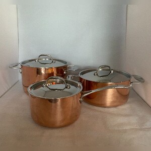 May include: Three copper pots with silver handles. The pots are nested inside each other, with the largest pot at the bottom and the smallest pot at the top.