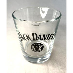 May include: A clear glass shot glass featuring the "Jack Daniel's" logo in black. The glass has a cylindrical form with a gently widening rim and a circular base. The "Old No. 7 Brand" logo is also displayed.