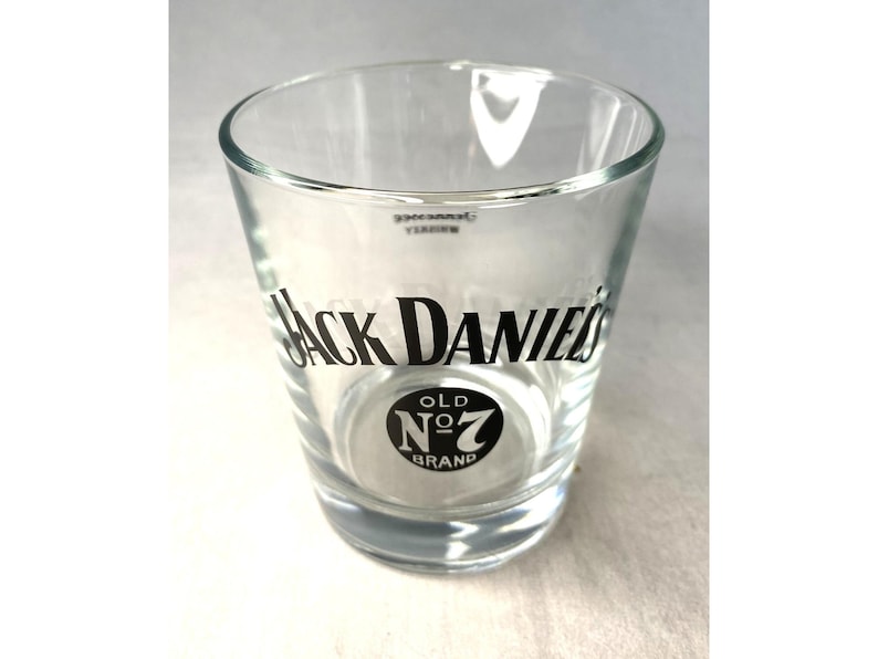 May include: A clear glass shot glass featuring the "Jack Daniel's" logo in black. The glass has a cylindrical form with a gently widening rim and a circular base. The "Old No. 7 Brand" logo is also displayed.
